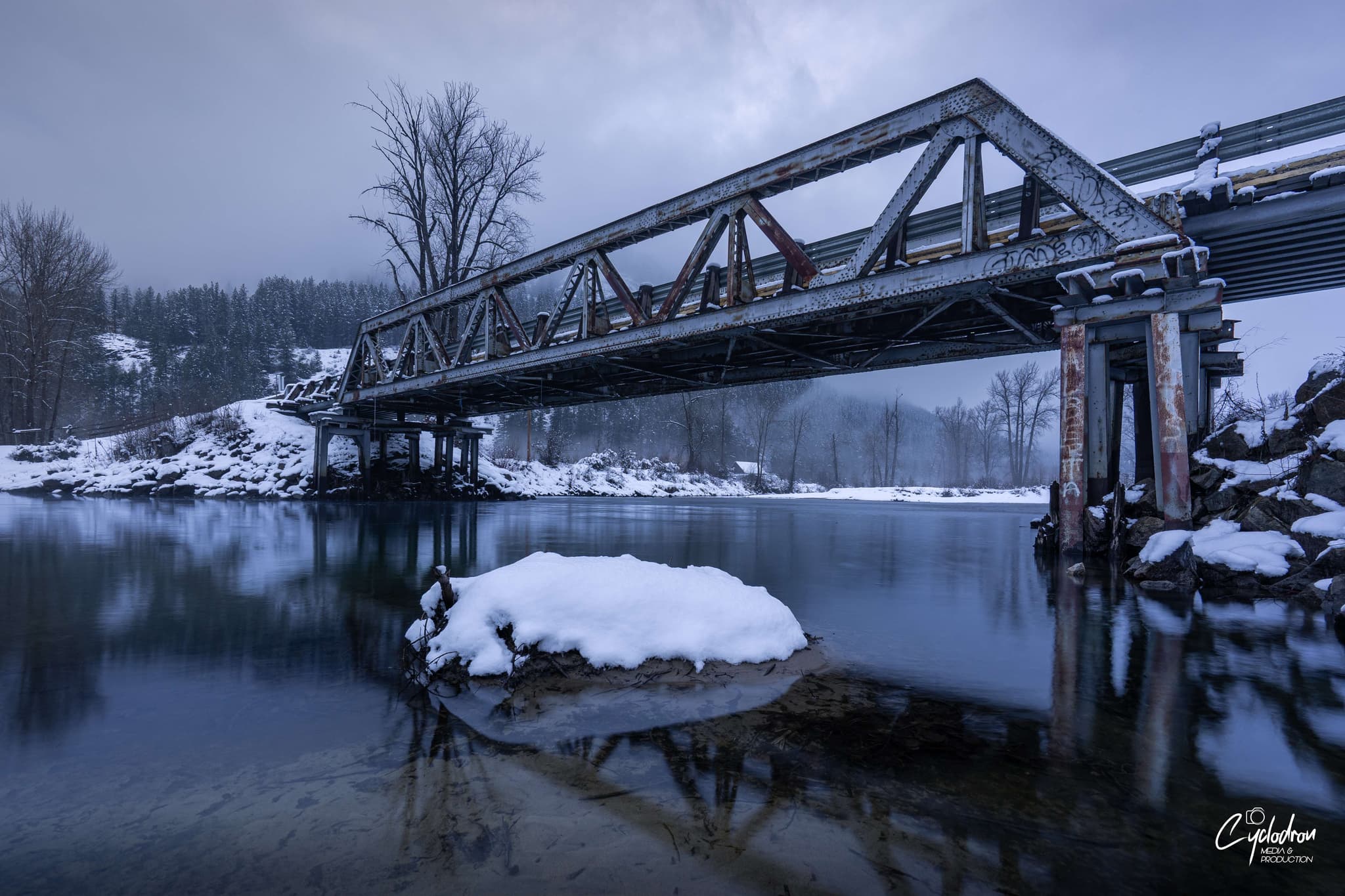 Historic metal truss bridge over calm river with snow-covered rocks in foreground during moody winter blue hour